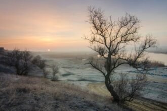 Havas táj napfelkeltével, fák és folyó látható a háttérben.