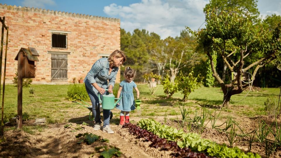 Egy nő és egy kislány közösen öntözik a kertben a zöldségeket, miközben a háttérben egy régi téglaépület látható.