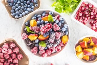 Tálban fagyasztott gyümölcsök, mint áfonya, málna és őszibarack, friss zöld levéllel díszítve.