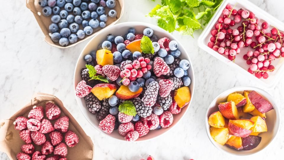 Tálban fagyasztott gyümölcsök, mint áfonya, málna és őszibarack, friss zöld levéllel díszítve.