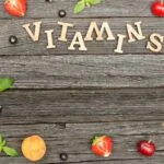 Friss gyümölcsök és zöld levelek egy fából készült asztalon, vitaminokkal kapcsolatos üzenettel.