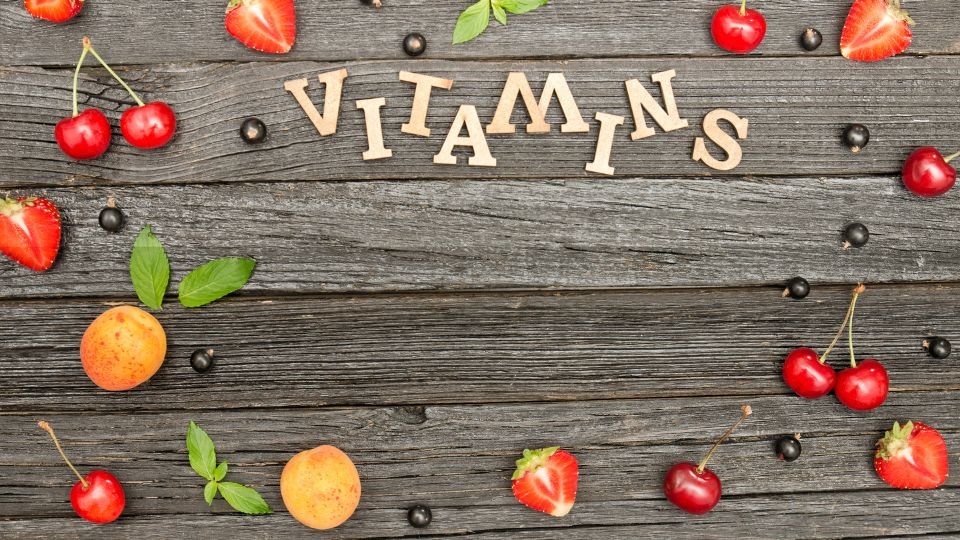 Friss gyümölcsök és zöld levelek egy fából készült asztalon, vitaminokkal kapcsolatos üzenettel.