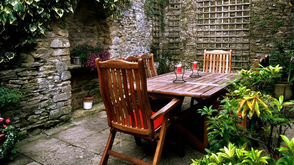 Egy hangulatos, helytakarékos kertasztal néhány székkel, mögötte kőfal és zöld növények.