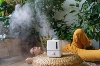 Egy fiatal nő relaxál a szobanövények között, miközben párásítót használ.