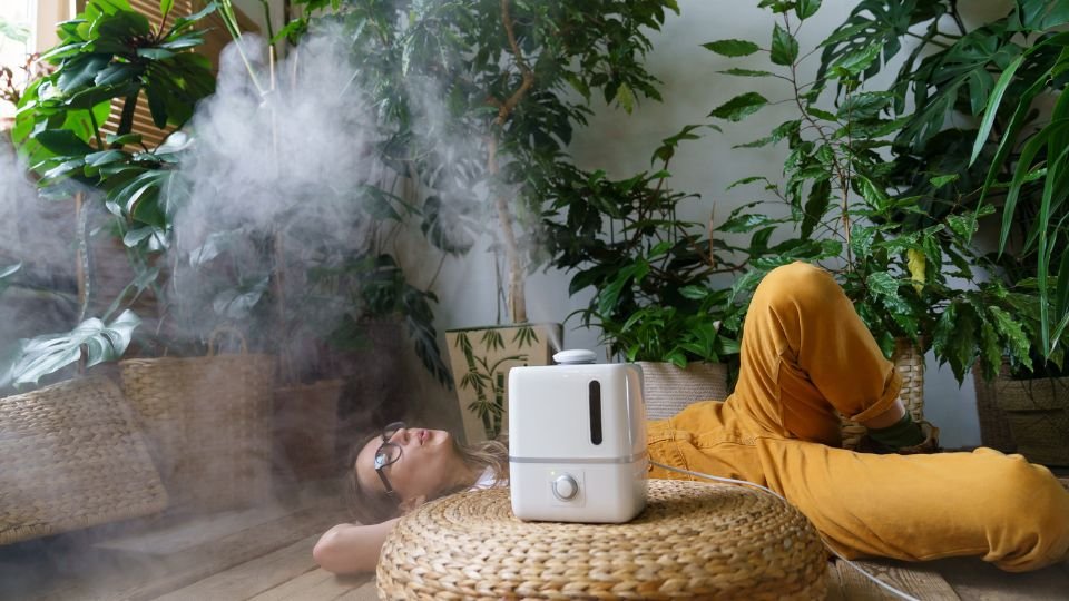 Egy fiatal nő relaxál a szobanövények között, miközben párásítót használ.