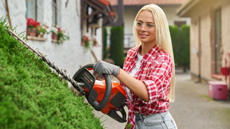 Egy fiatal nő sövényvágóval formázza a zöld sövényt a kertben, vidám hangulatban.