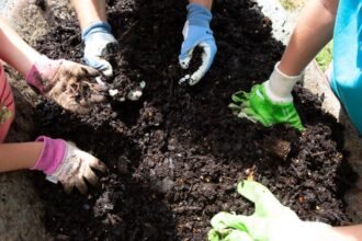 Gyerekek és felnőttek komposztot készítenek, kezeik a földben, vidám kertészkedés.
