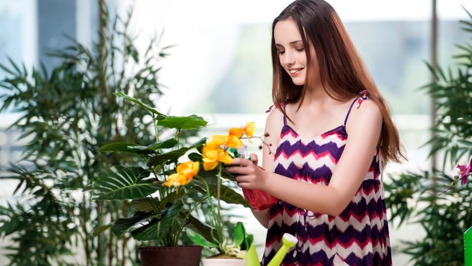 Egy fiatal nő gondozza a virágait egy világos, zöld növényekkel teli szobában.