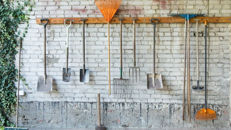 Kerti szerszámok rendezett tárolása egy téglafalon, zöld növények mellett.