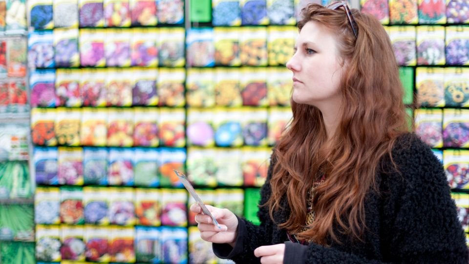 Egy fiatal nő vetőmagokat nézeget a boltban, háttérben színes csomagolásokkal.