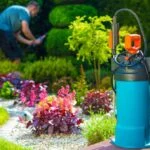 Kertész permetező spray a gyönyörű kertben, virágokkal és zöld növényekkel.