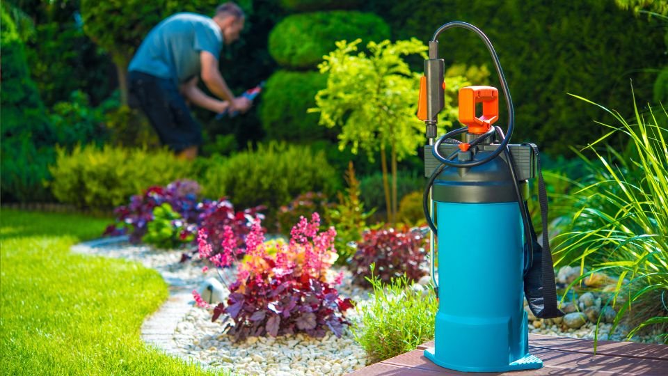 Kertész permetező spray a gyönyörű kertben, virágokkal és zöld növényekkel.