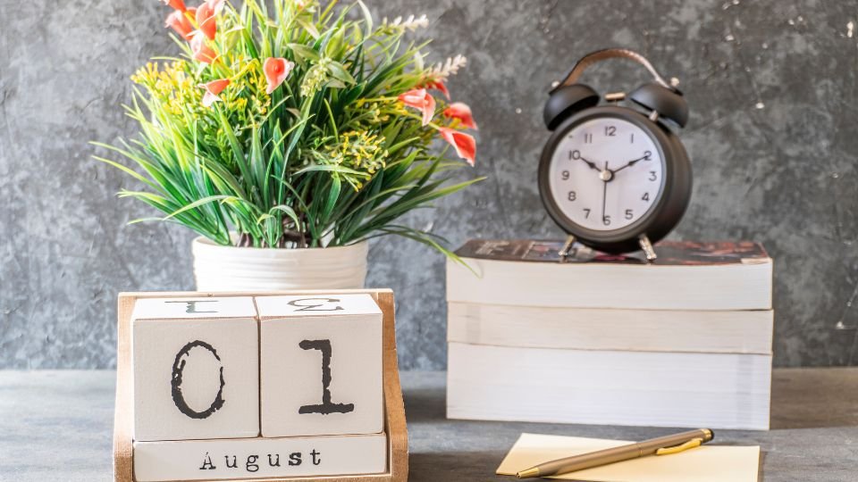 Augusztusi naptár, virágcserép és óra egy íróasztalon.