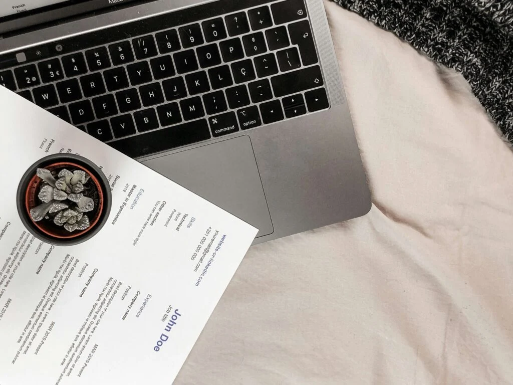 Egy laptop, mellette egy kis cserép növény és egy önéletrajz, barátságos környezetben.