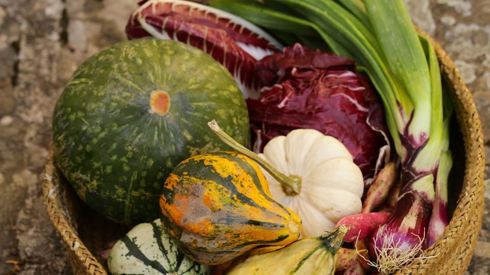 Különböző őszi zöldségek és fűszernövények egy kosárban, köztük tökök, póréhagyma és saláták.