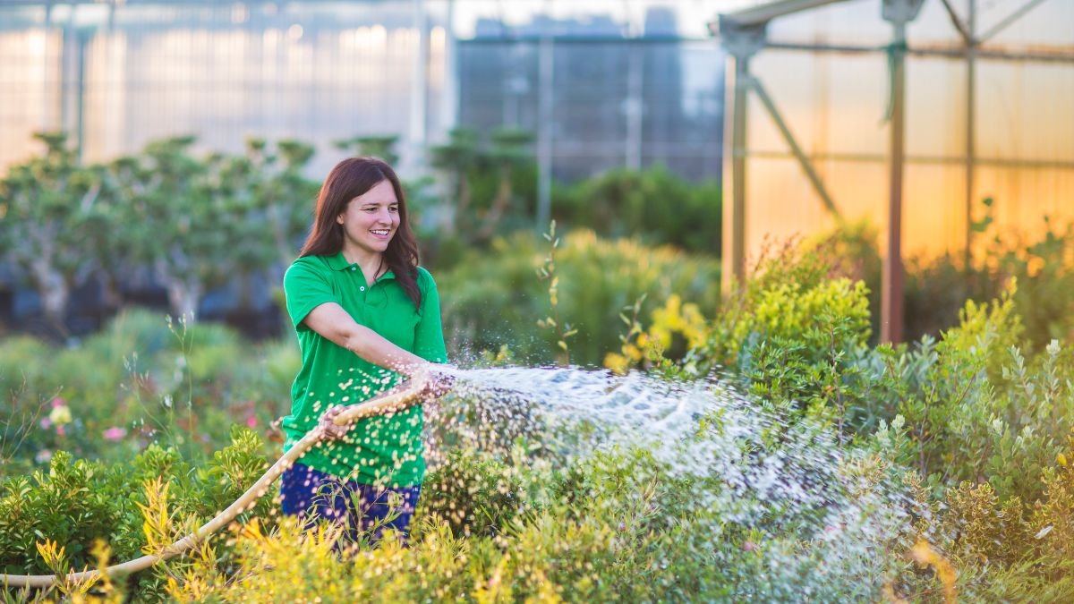 Egy nő locsolja a kertet, miközben zöld növények között áll, mosolyogva.
