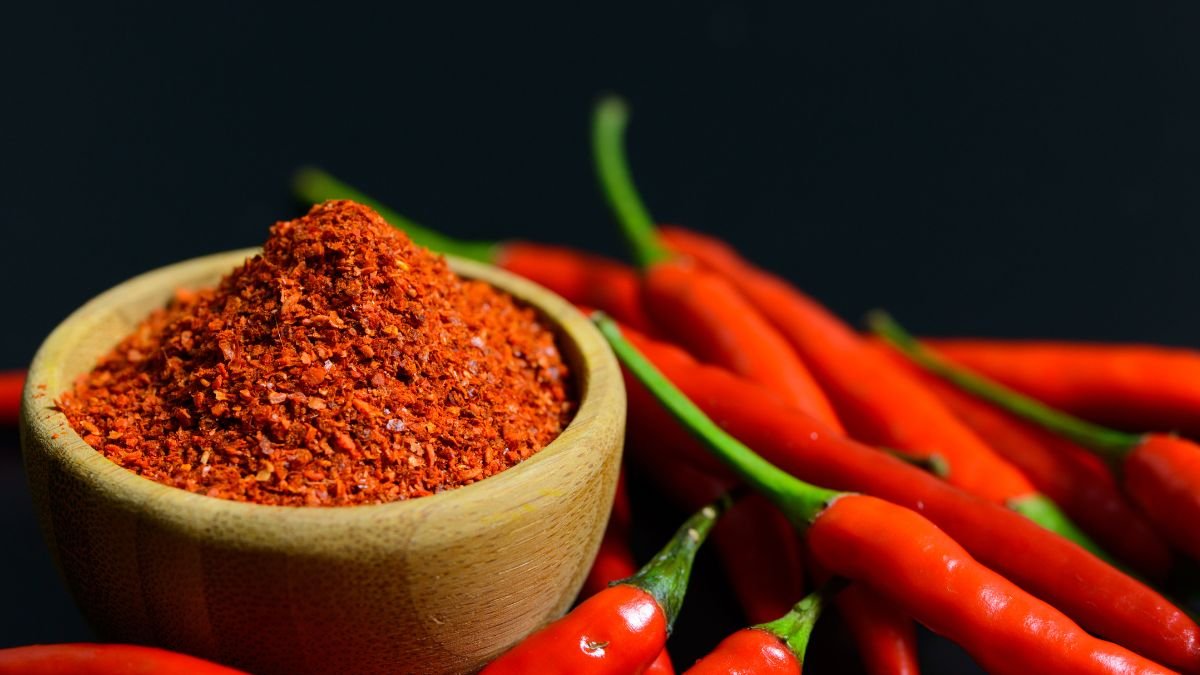 Egy fából készült tálban chili paprika por található, körülötte friss, piros chili paprikák helyezkednek el.